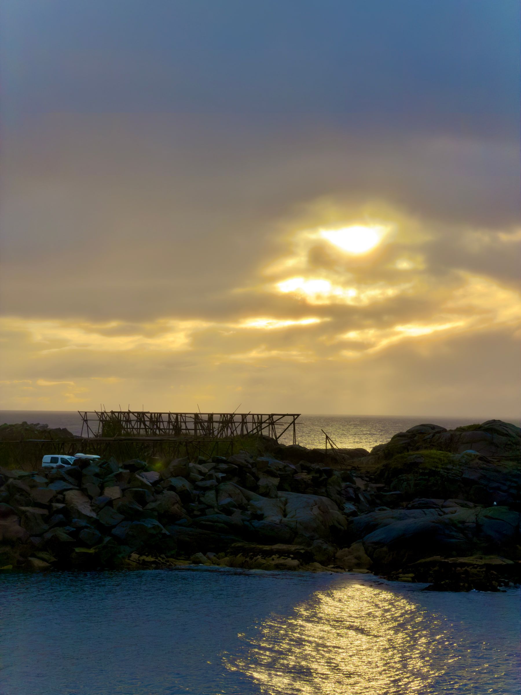 Empty wooden stockfish drying racks silhouetted on a rocky shore, with low sun breaking through cloud and laying gold on the sea