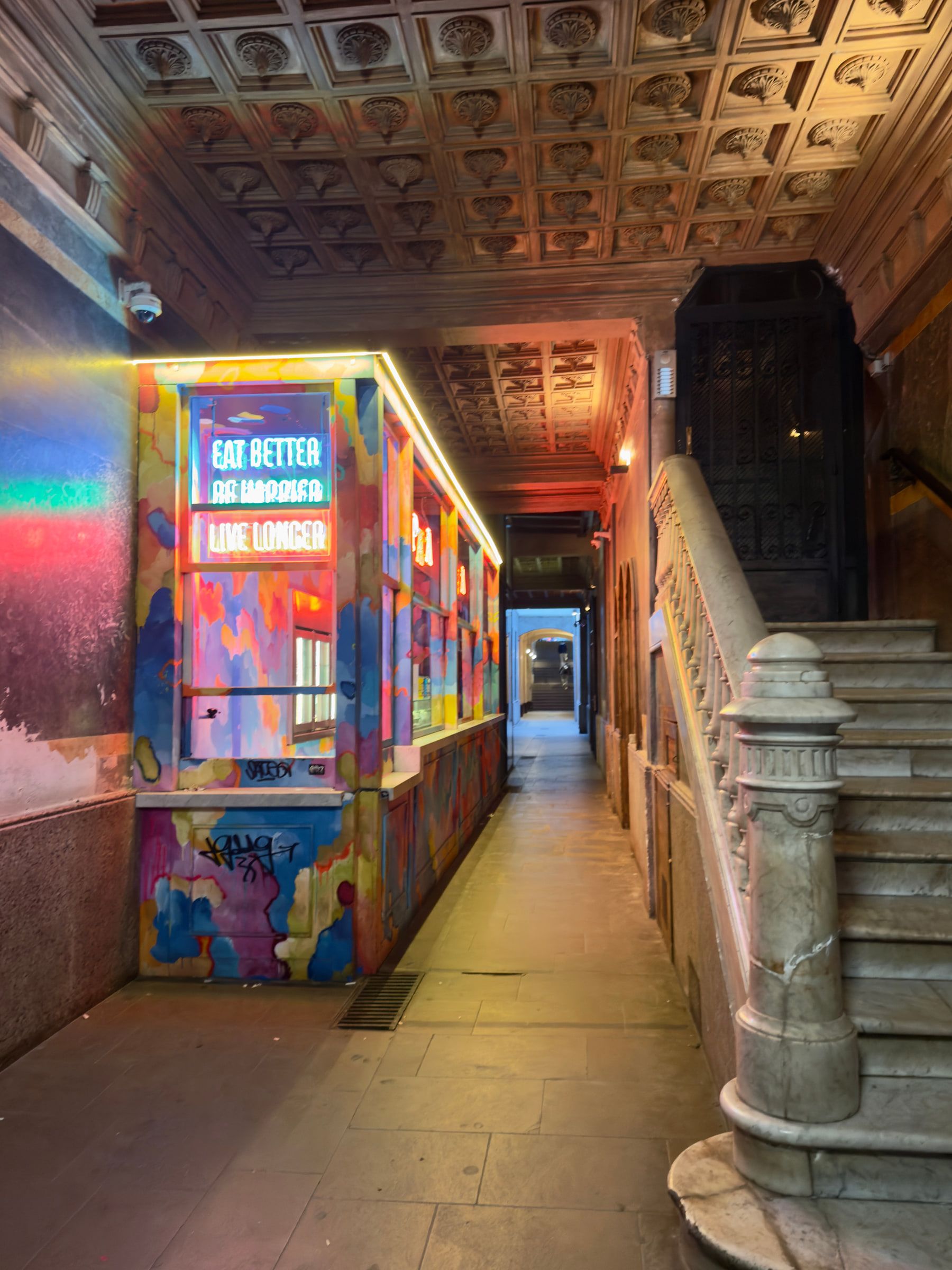 A corridor with an ornate coffered ceiling and a long mural in bright blues and pinks, a neon sign reading eat better live longer