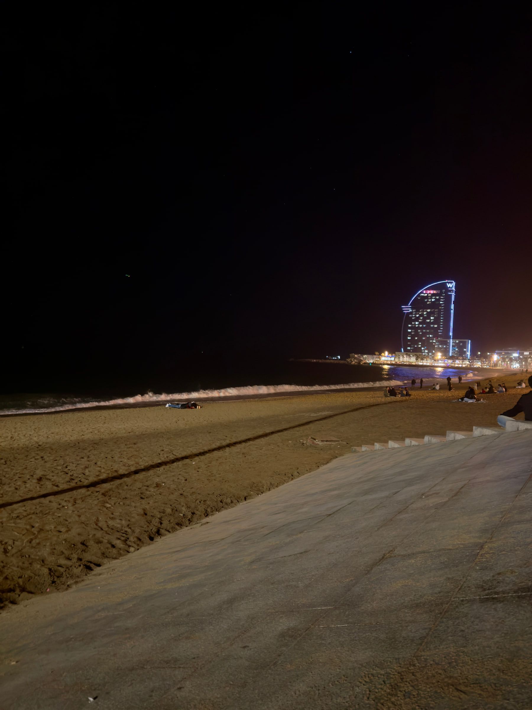 A dark empty beach at night, the lit sail-shaped silhouette of a seafront hotel standing at the far end of the sand