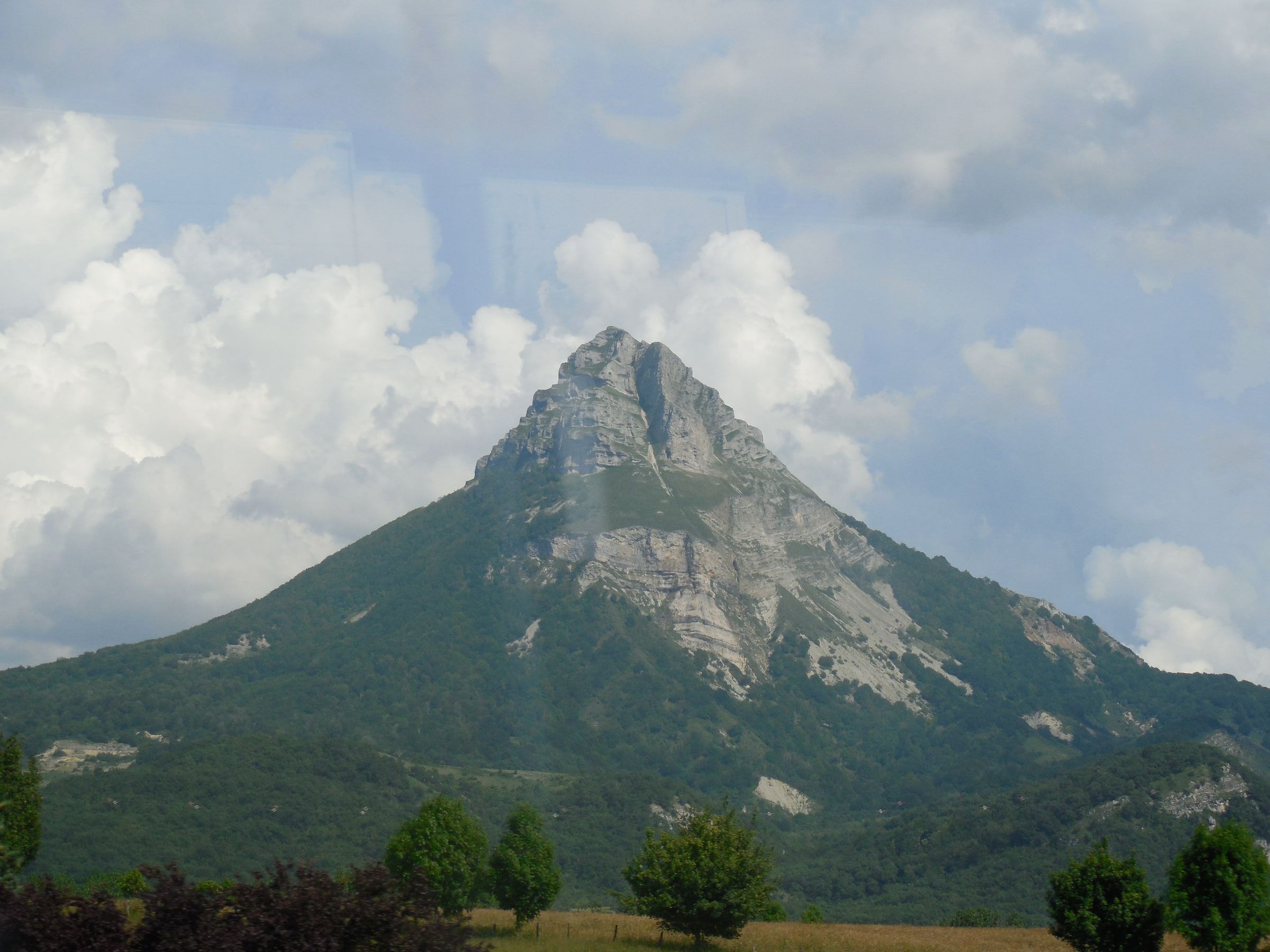 A steep conical mountain rising out of green farmland under heavy white cloud