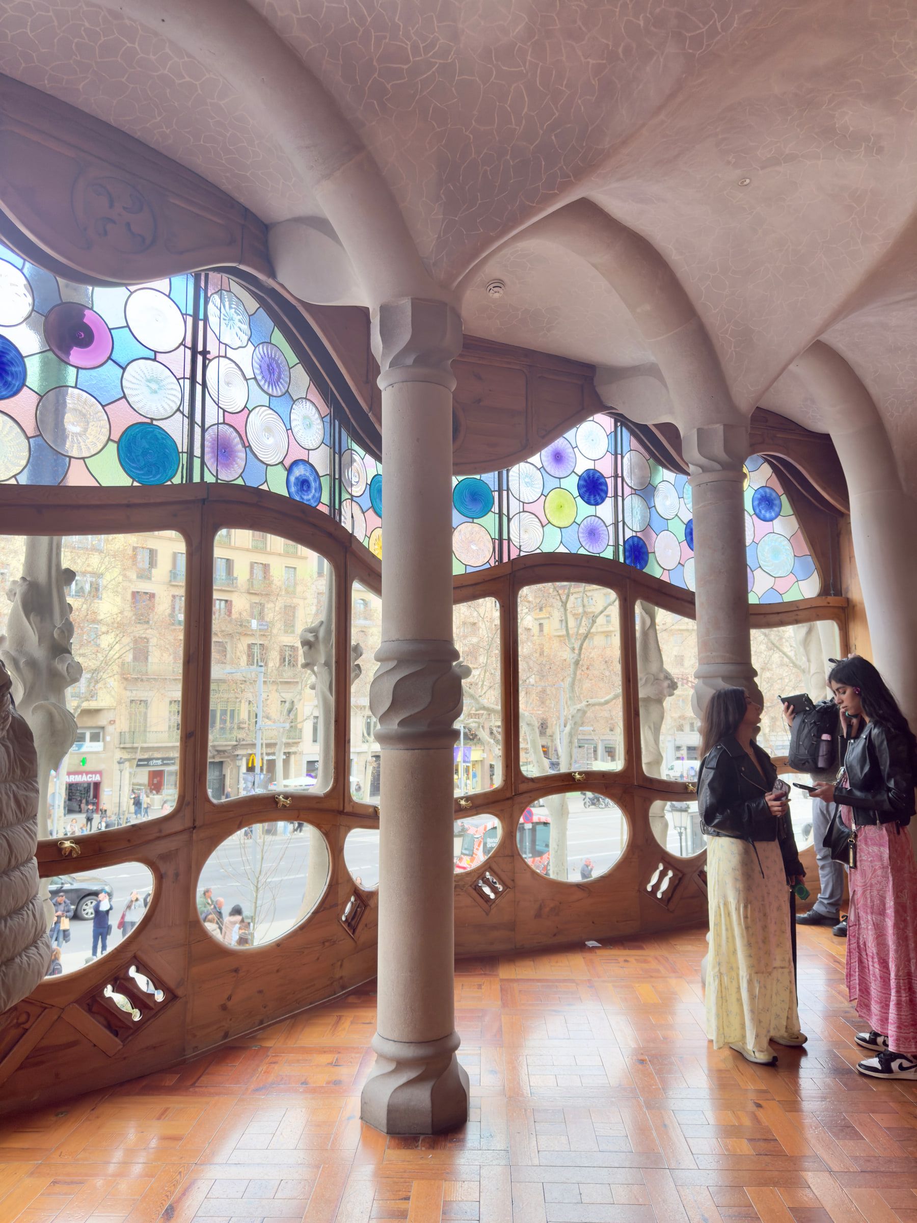 A room with bone-like stone columns and a wall of wavy glazing set with blue circular panes, visitors standing talking at the windows