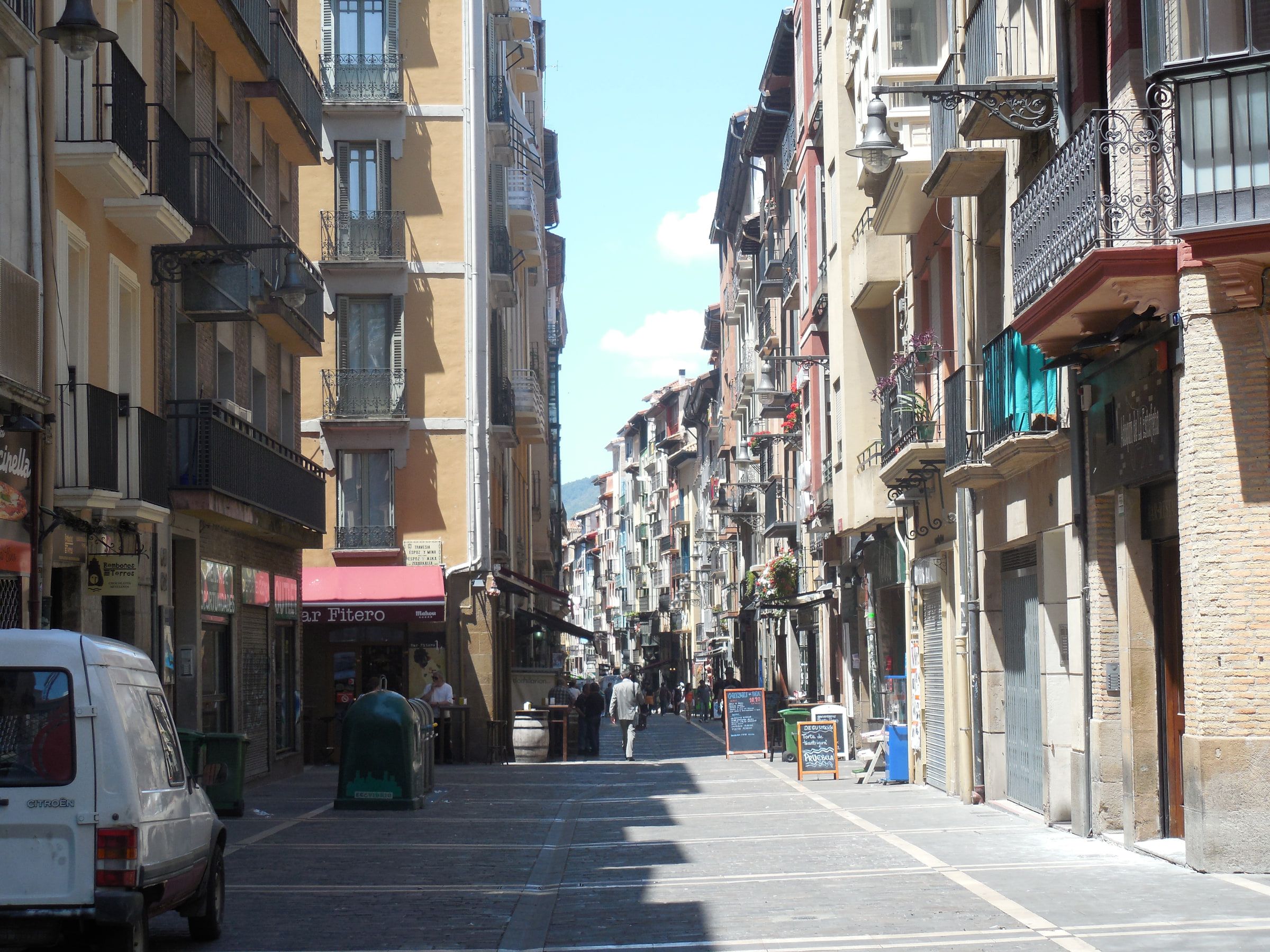 A narrow old-town street of balconied buildings in bright sun, a van parked at the near end