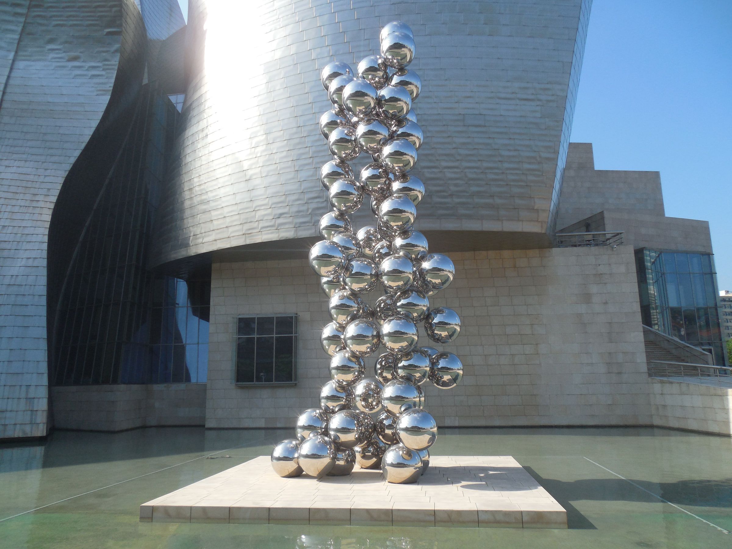 A tall column of mirrored steel spheres standing before a titanium-clad museum wall