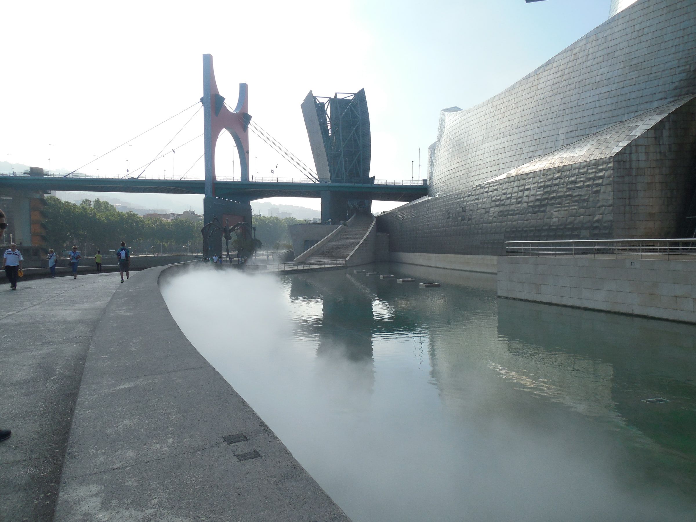 A red-pylon bridge passing over a titanium-clad museum beside a river, mist lying on the water