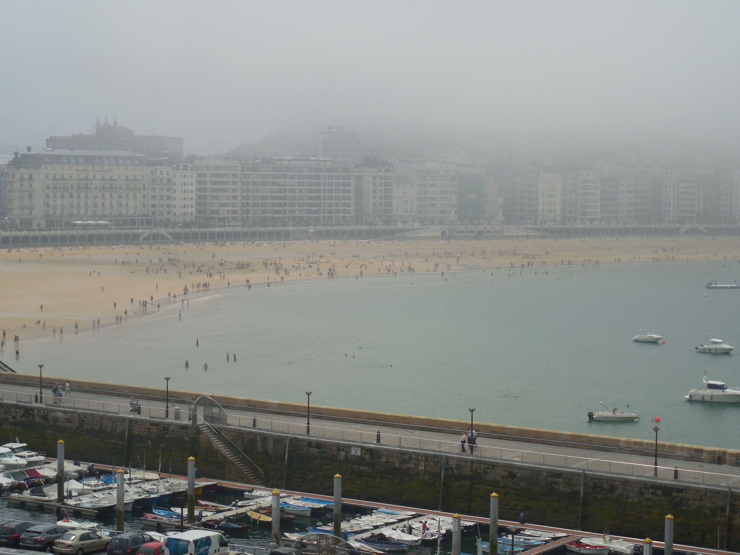 A crescent beach and bay under heavy fog, a marina in the foreground and buildings faint behind