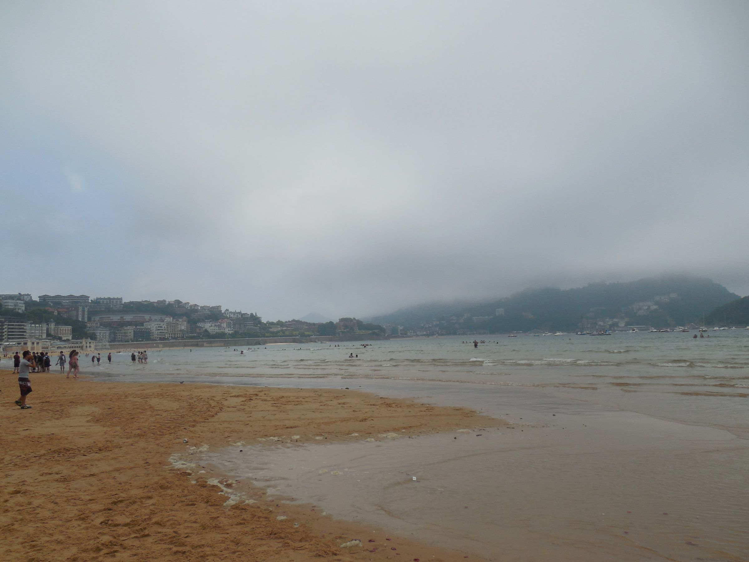 A broad beach at low tide under grey cloud, a wooded headland standing behind the town