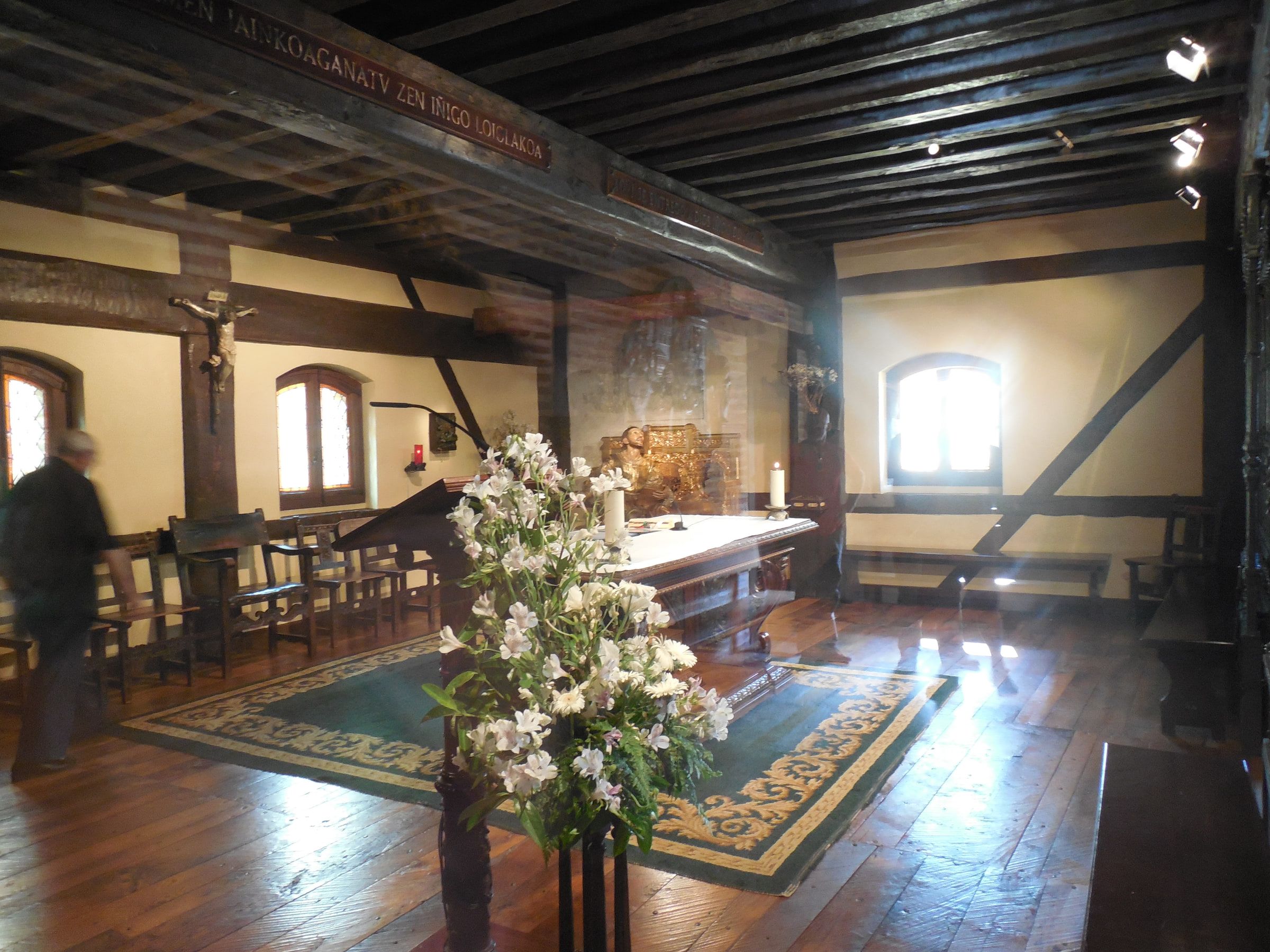 A chapel interior with heavy timber beams, an altar dressed in white flowers, a Basque inscription running along the wall