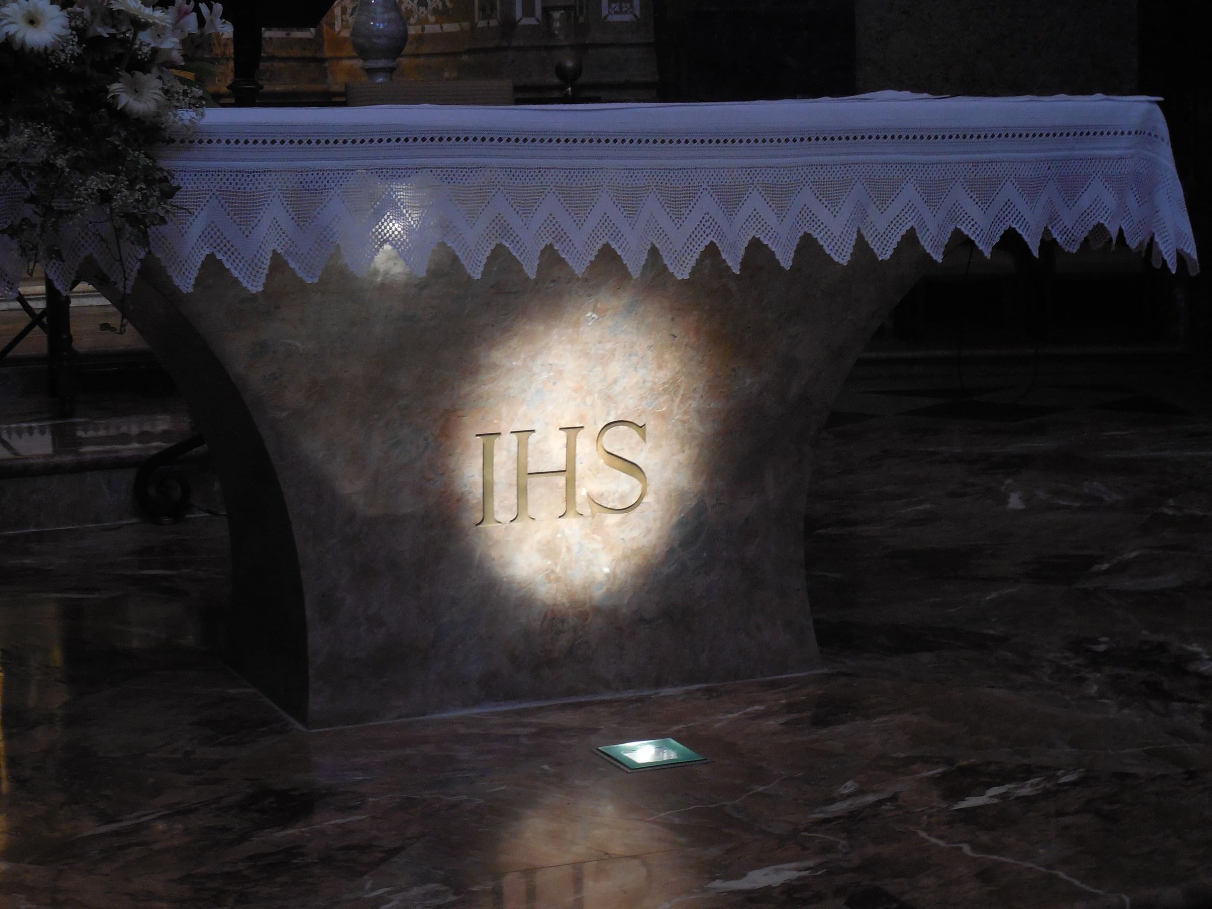 A dark stone altar lit from above, an IHS monogram carved into its face beneath a white lace cloth