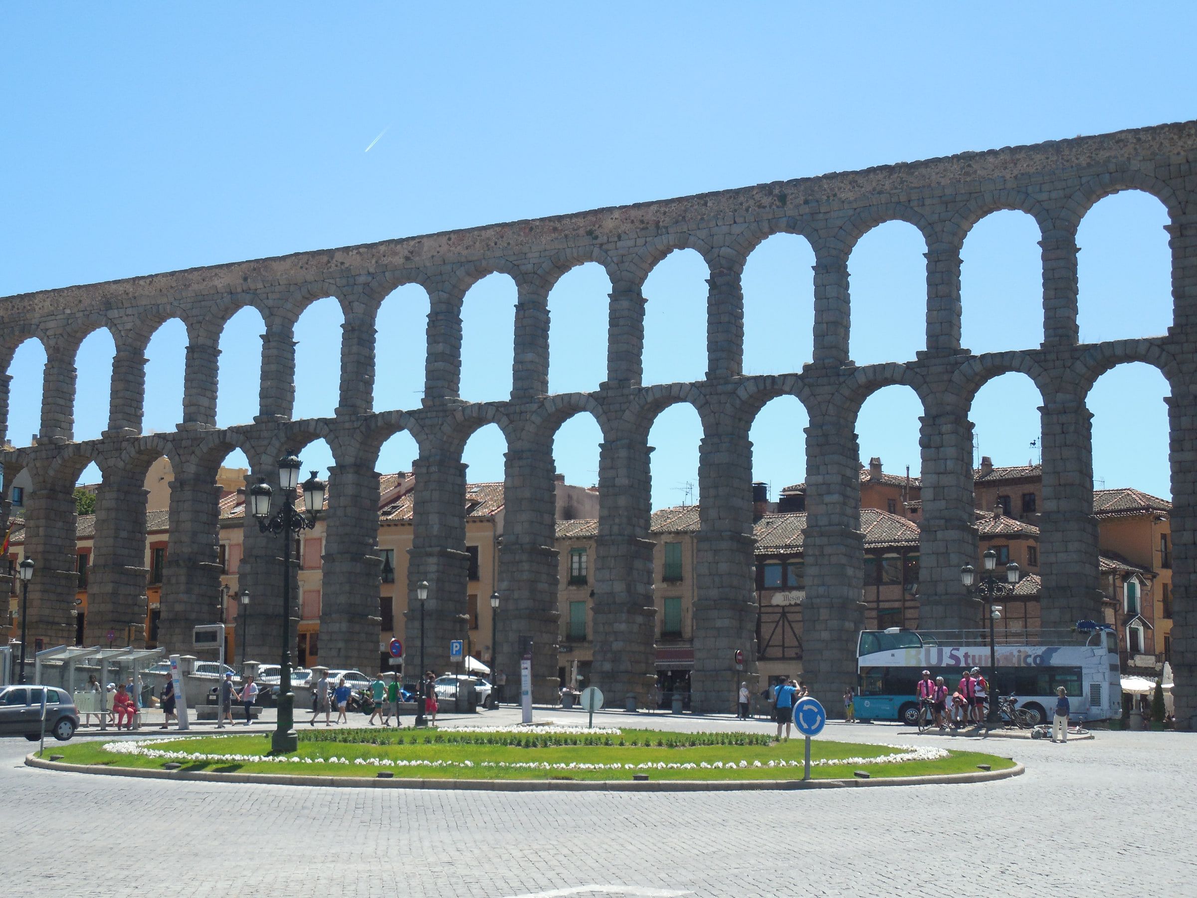 A Roman aqueduct of two tiers of arches crossing behind a town square and a planted roundabout