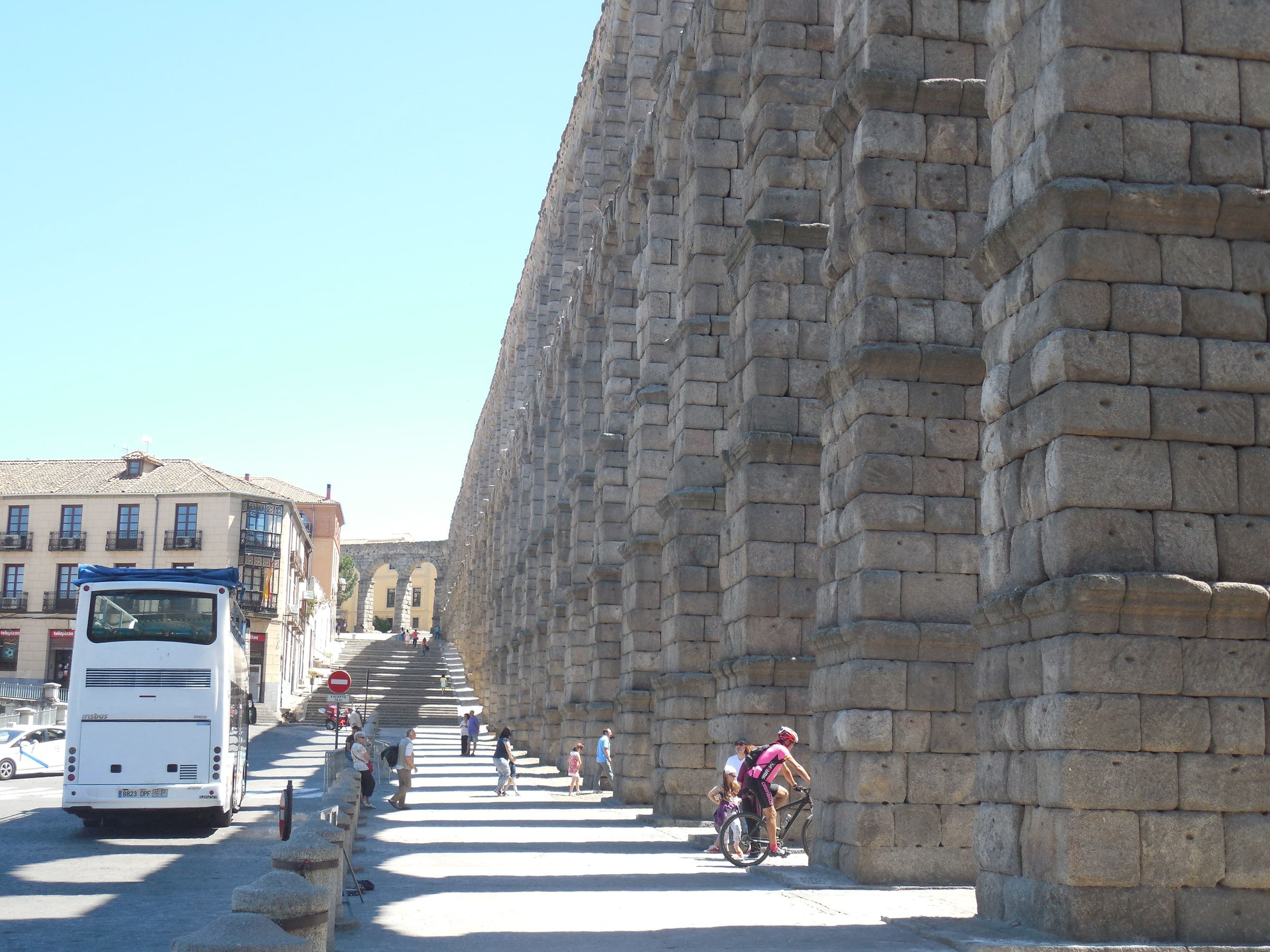 The massive granite piers of a Roman aqueduct seen close along a street, a coach parked beneath