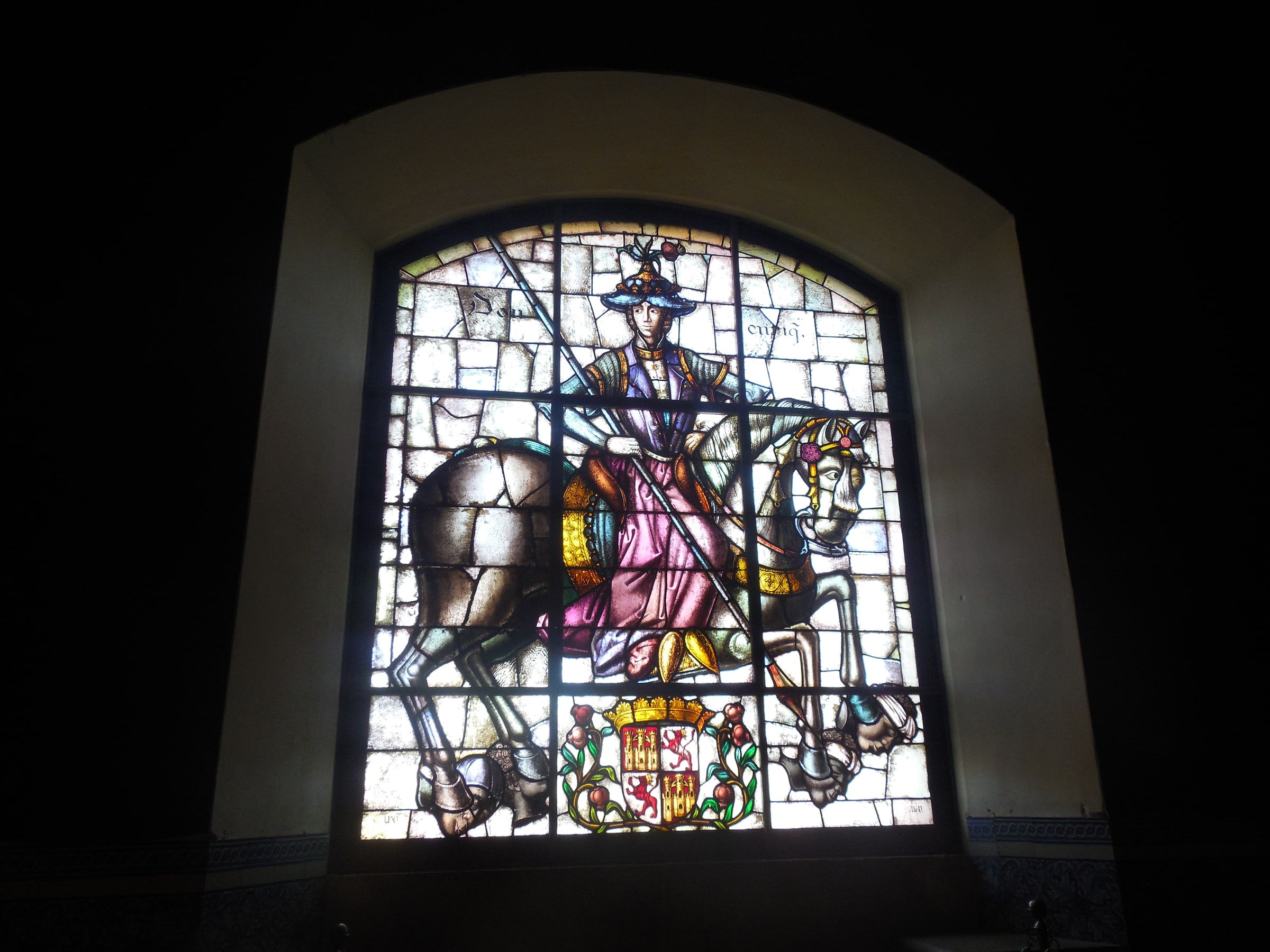 A stained-glass window of an armoured rider on horseback above a coat of arms