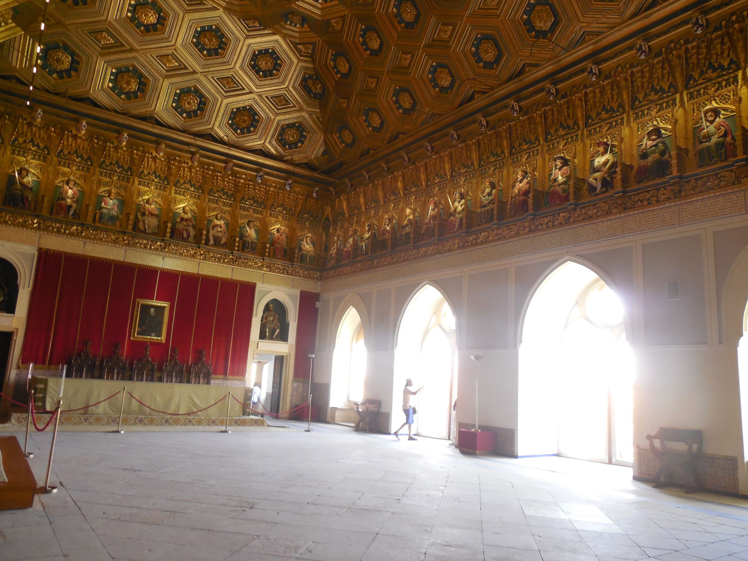 A long hall with a gilded coffered ceiling above red-hung walls and tall arched windows