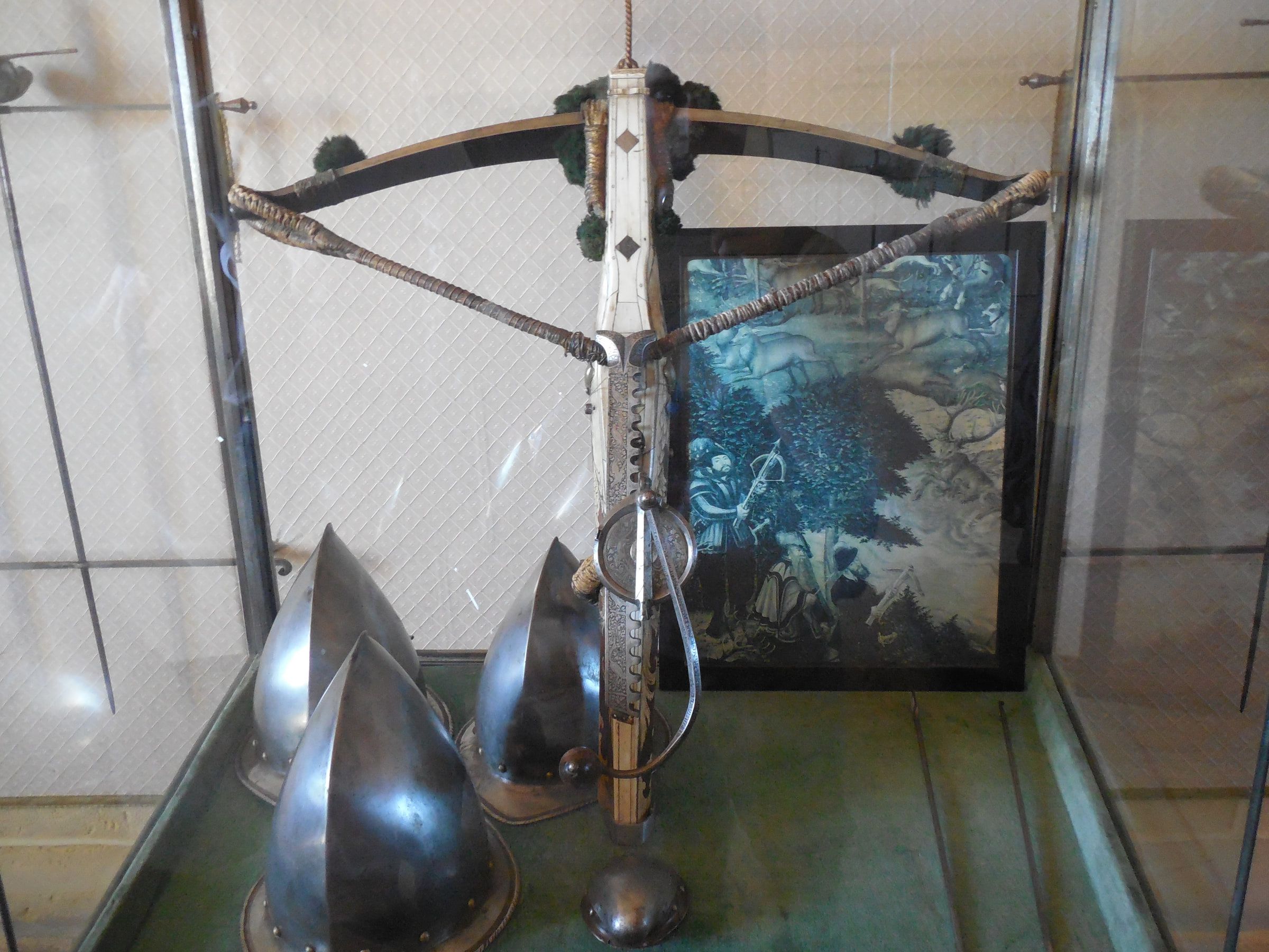 A crossbow and steel helmets displayed together in a glass museum case