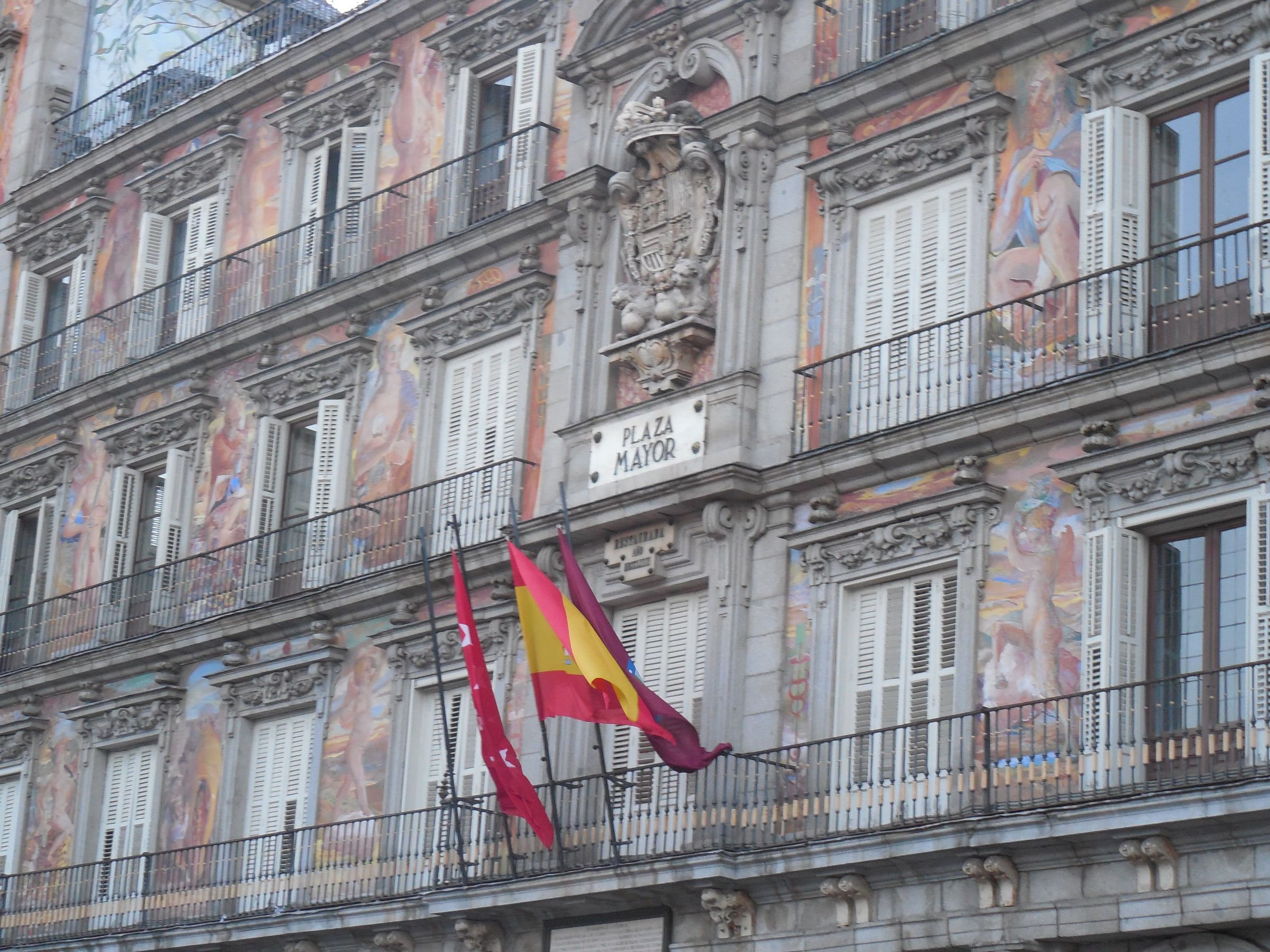 A grand square's painted facade, frescoed figures between balconied windows, flags hanging below