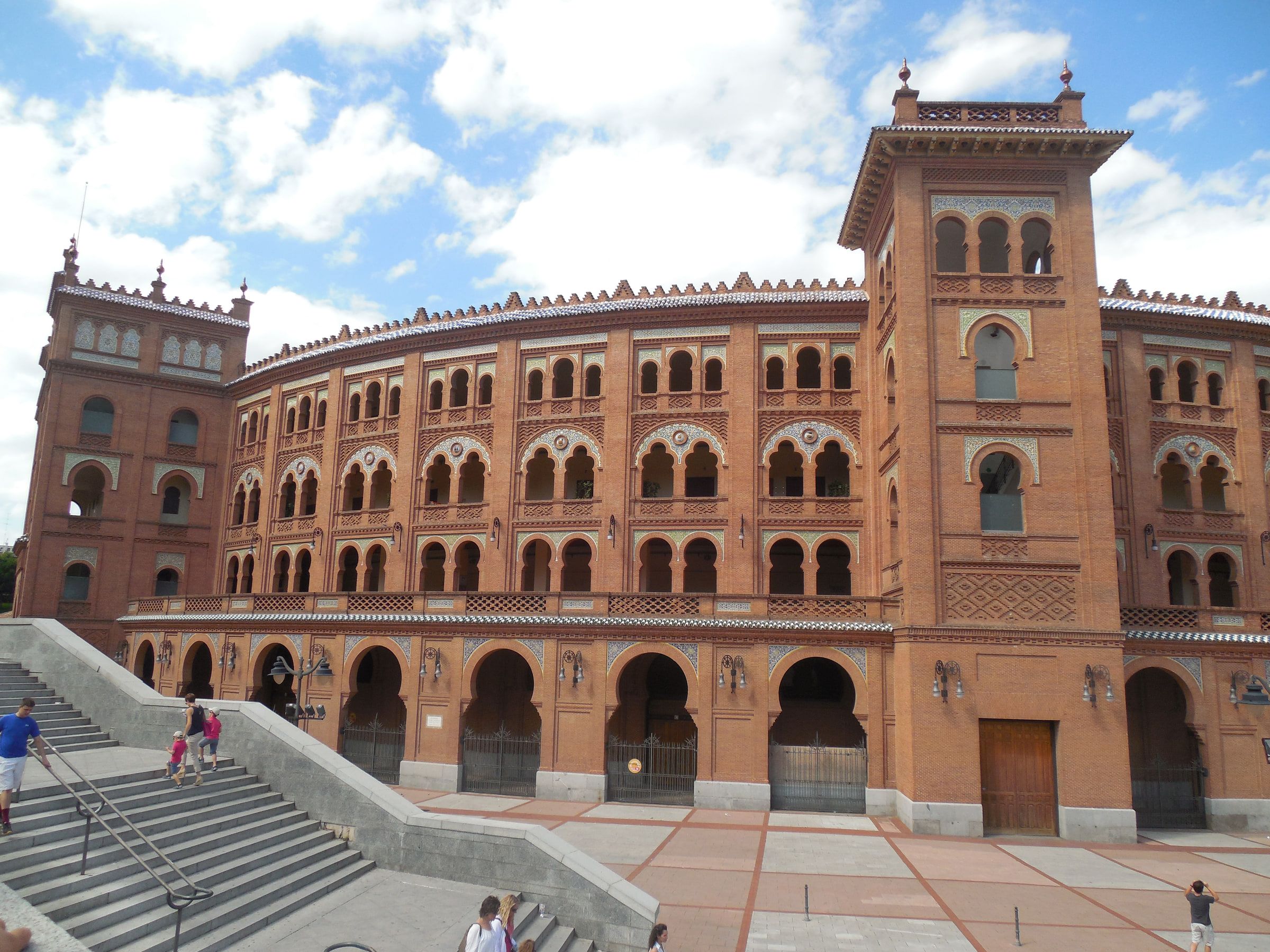 A large brick bullring with horseshoe arches and tiled decoration, steps rising to its entrance