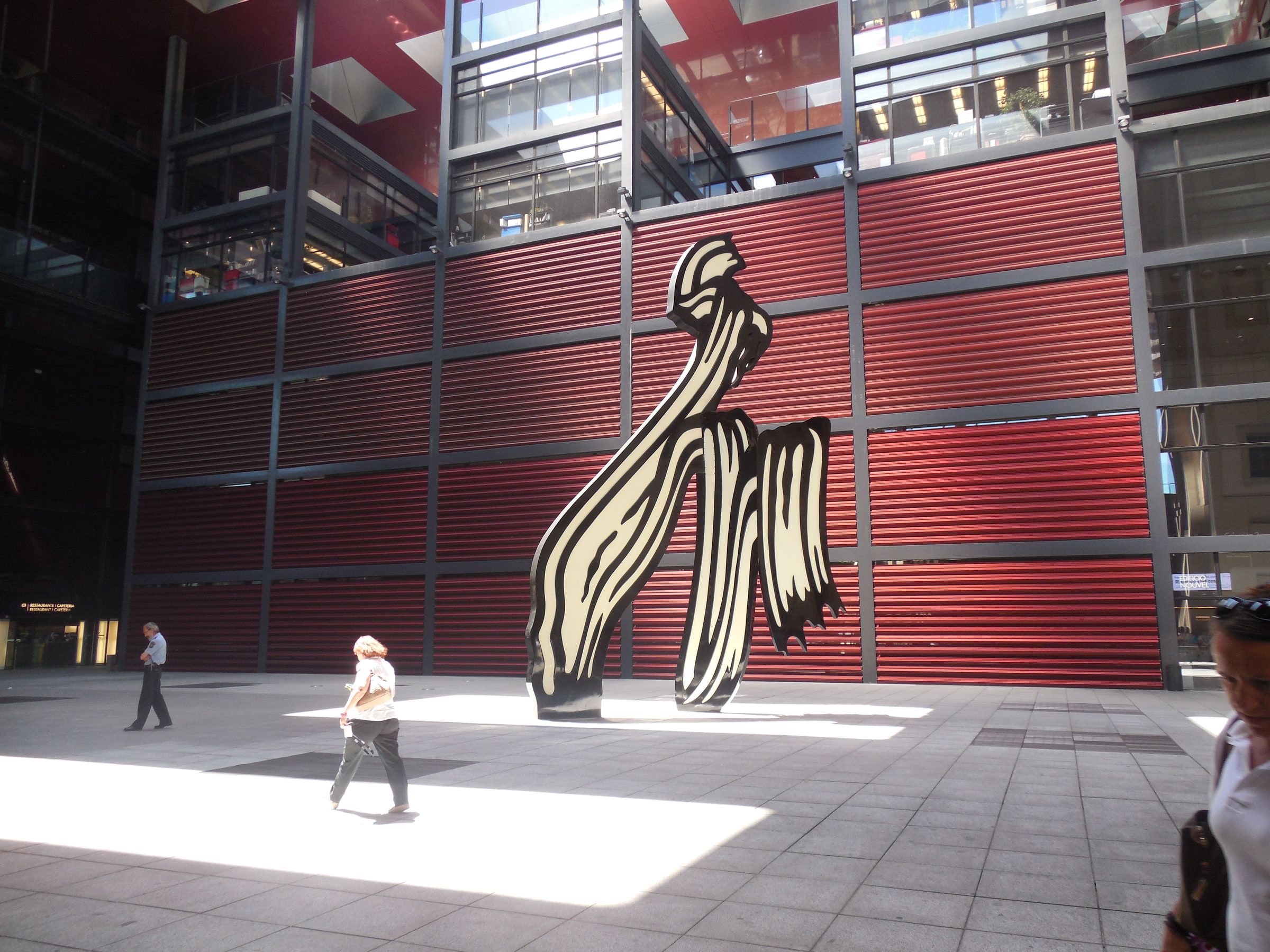 A tall sculpture of a folded brushstroke in black and yellow standing before a red-panelled modern building