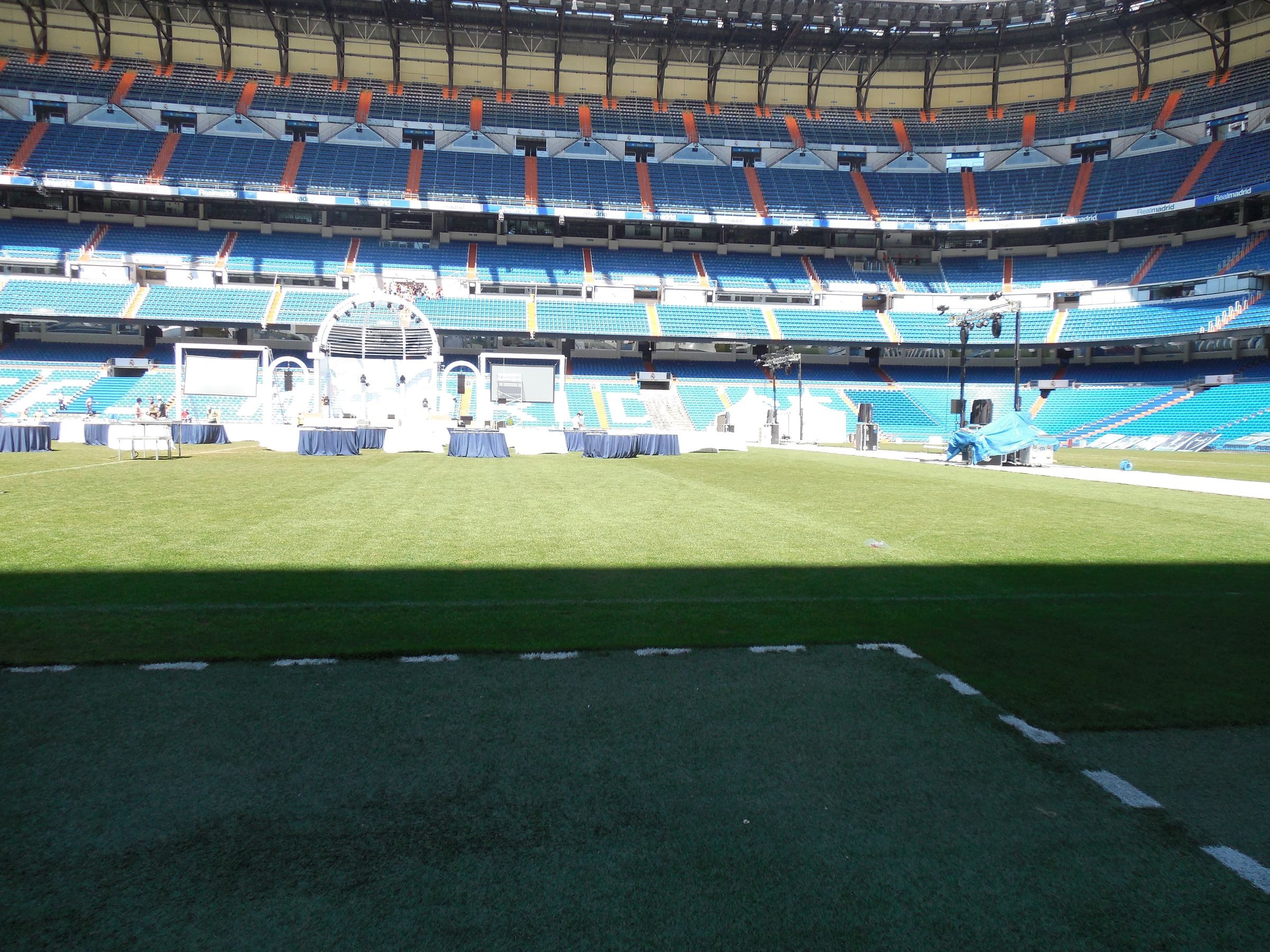 A football pitch from ground level, stands rising behind it and staging on the far side
