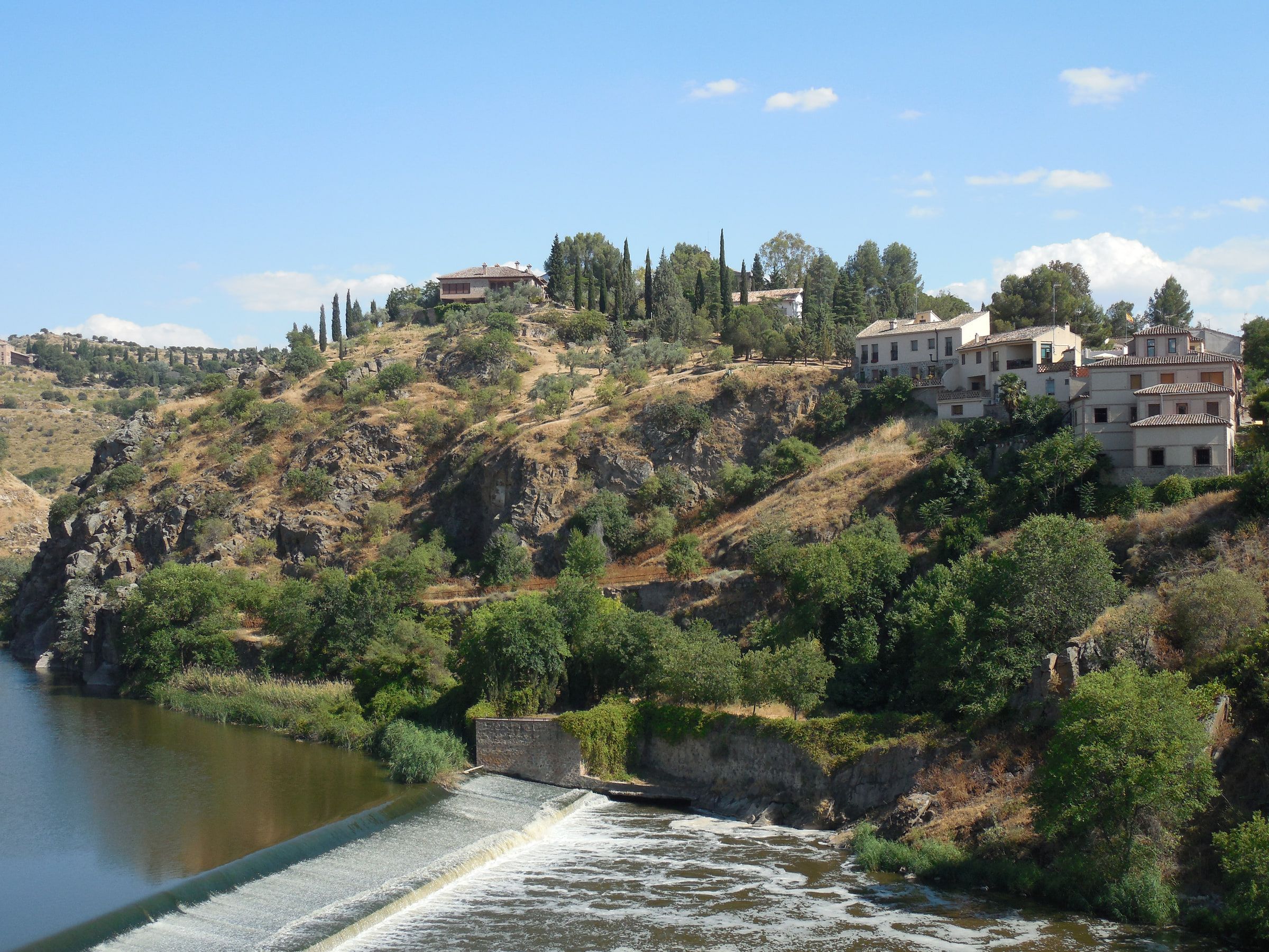 A weir crossing a river below a rocky slope topped with houses and cypresses