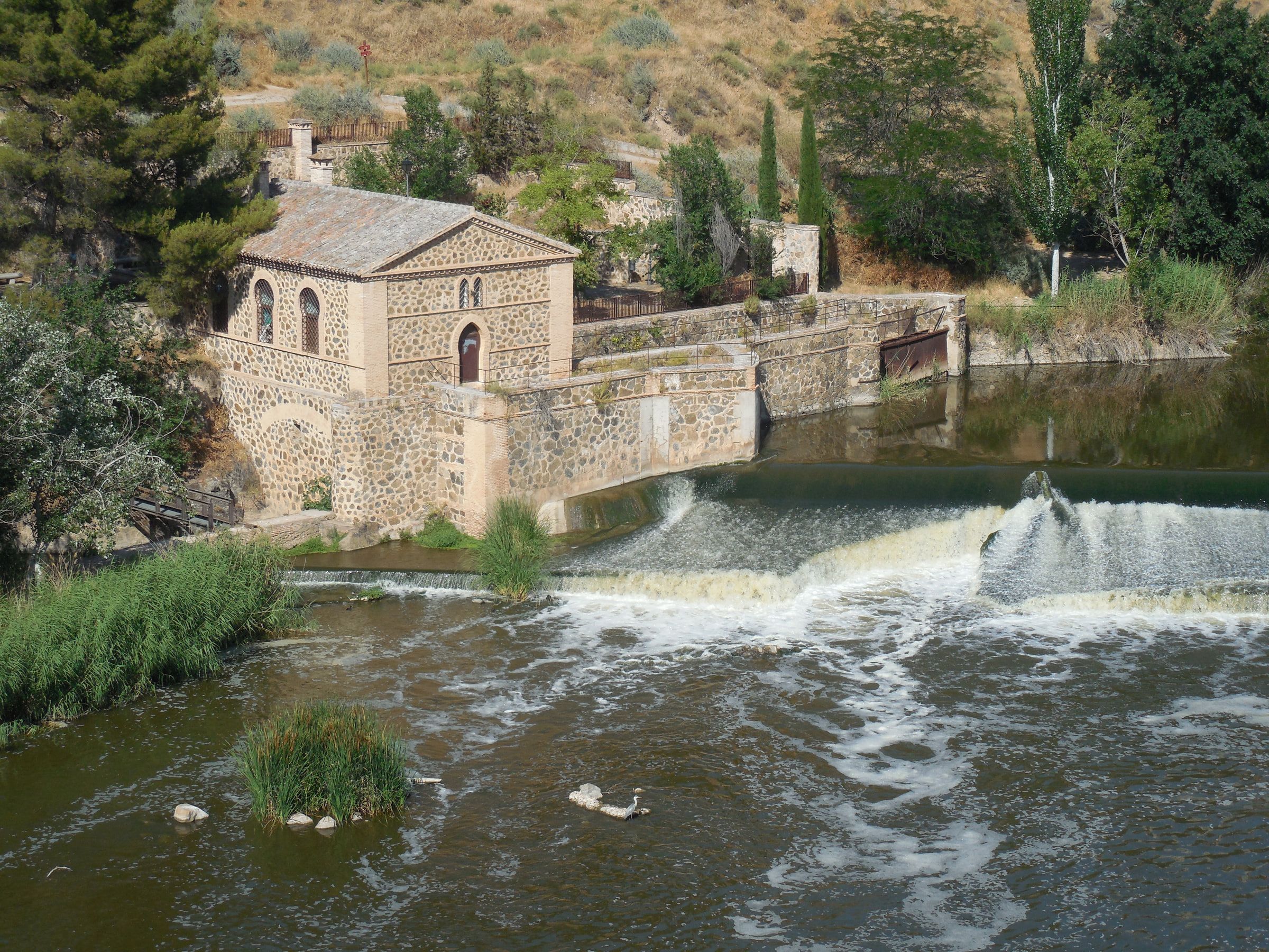 A stone building with arched windows standing at the end of a weir, water breaking white beneath it